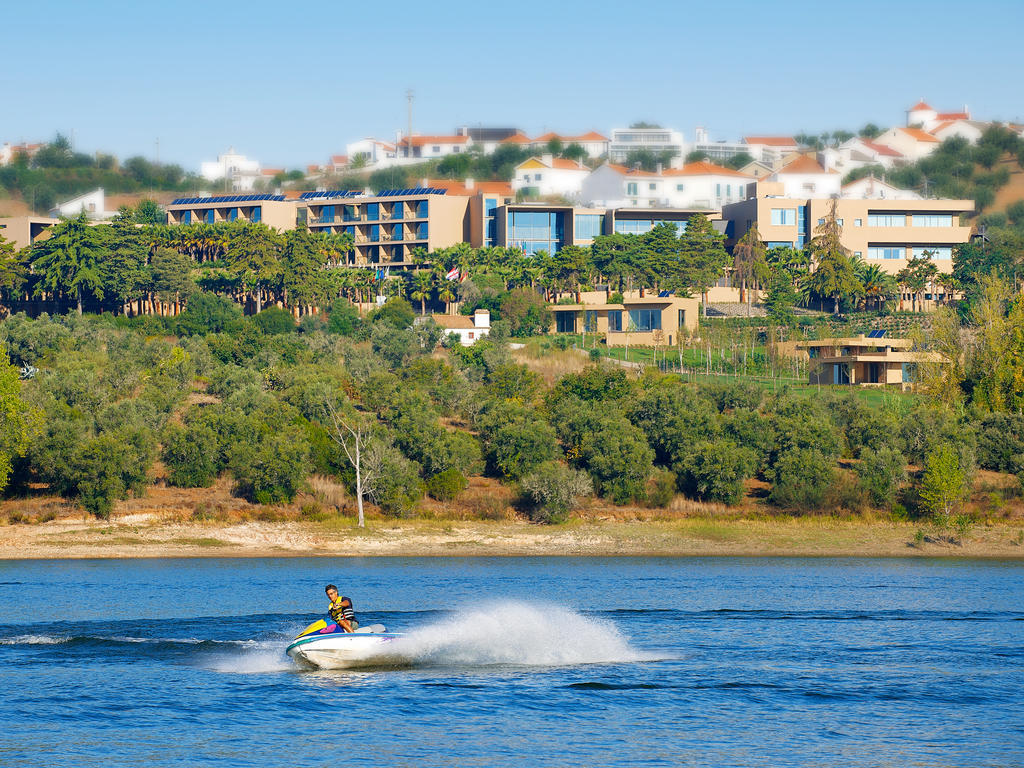 Ap Lago Montargil - Conference & Spa Εξωτερικό φωτογραφία The photo depicts a person riding a jet ski on a body of water. In the background, there are modern buildings and lush greenery, suggesting a scenic landscape, likely near a resort or upscale residential area. The clear blue sky and calm water enhanc