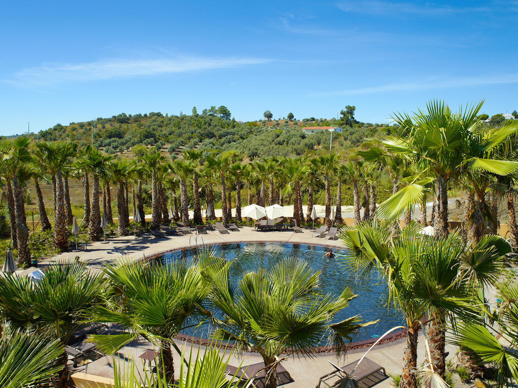 Ap Lago Montargil - Conference & Spa Εξωτερικό φωτογραφία The photo depicts a beautiful outdoor area featuring a circular swimming pool surrounded by palm trees. The pool area has several lounge chairs arranged around it. In the background, there is a lush green hillside, and the sky is clear with a bright 