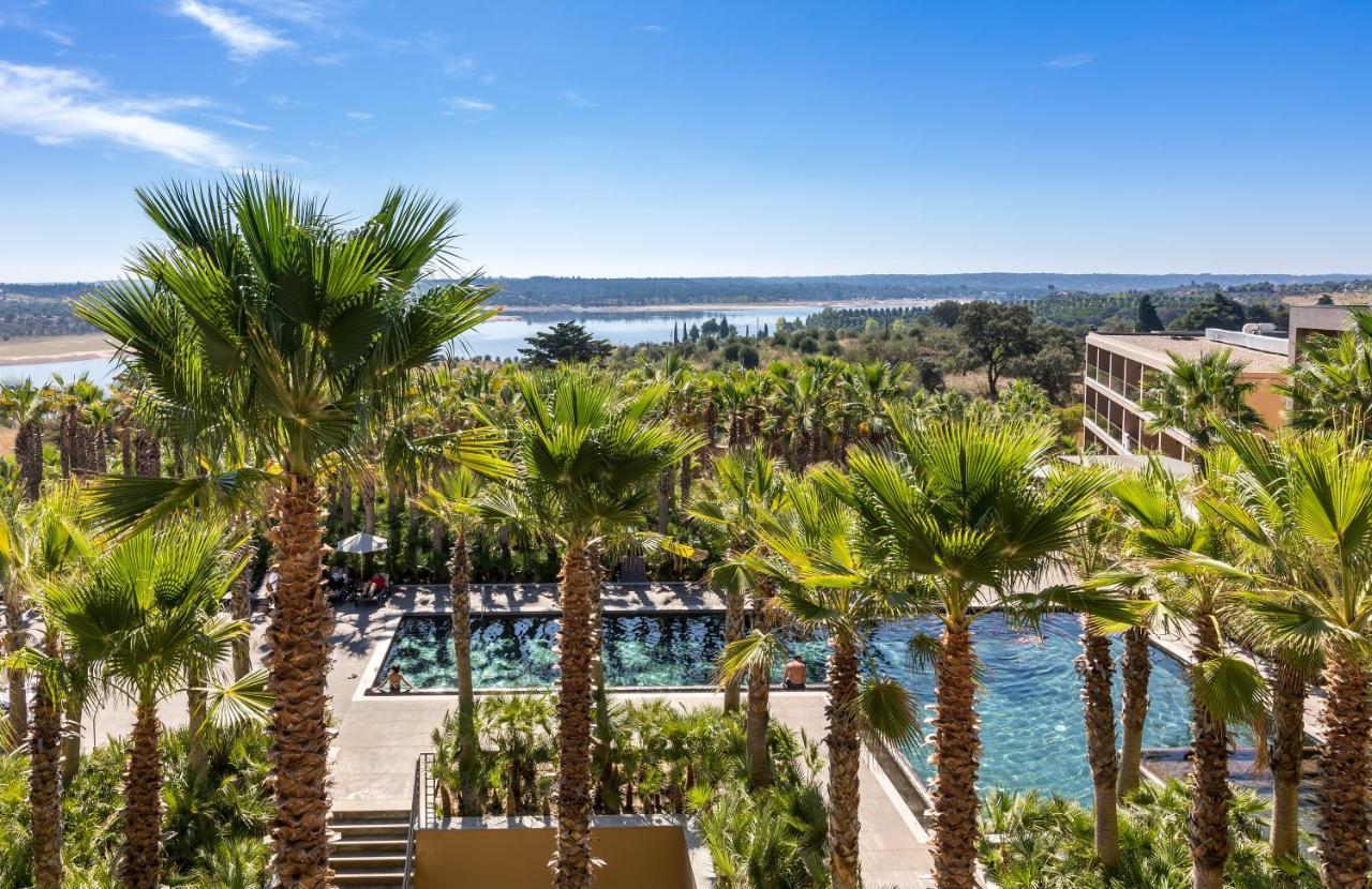 Ap Lago Montargil - Conference & Spa Εξωτερικό φωτογραφία The photo depicts a picturesque resort landscape with a clear blue sky. In the foreground, there are tall palm trees surrounding a swimming pool area. The swimming pool has a modern design and is surrounded by lush greenery. In the background, you ca
