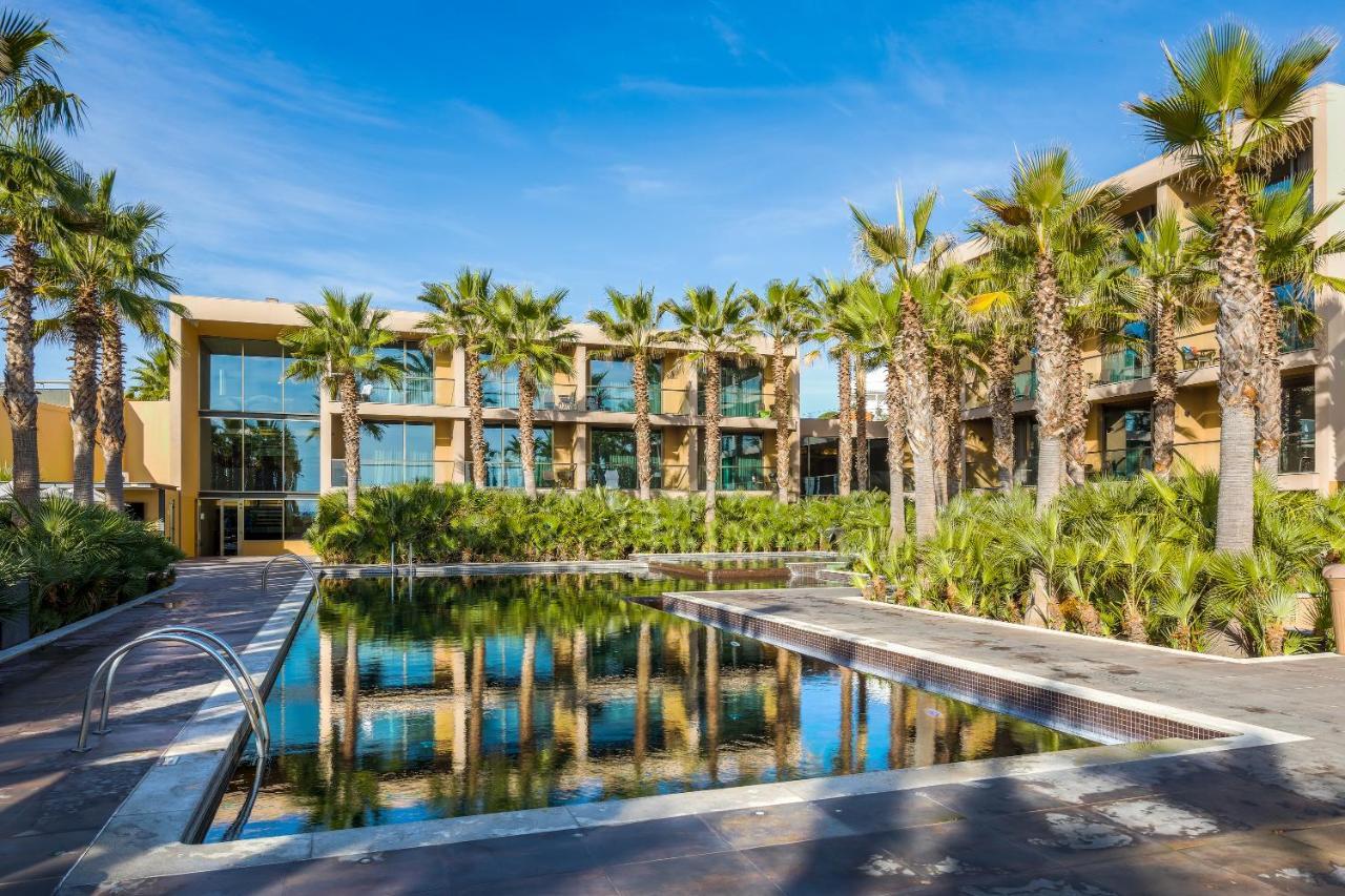 Ap Lago Montargil - Conference & Spa Εξωτερικό φωτογραφία The photo features a modern building surrounded by lush greenery and palm trees. In the foreground, there is a serene swimming pool with a reflective surface that captures the surrounding environment. The architecture of the building appears to have 