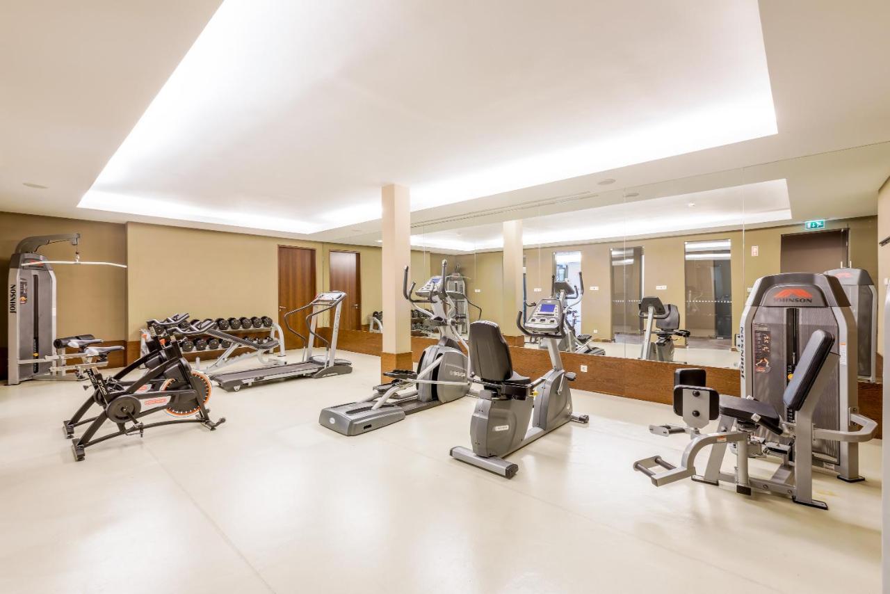 Ap Lago Montargil - Conference & Spa Εξωτερικό φωτογραφία The image shows a modern gym interior featuring various exercise equipment. On the left, there are free weights and weight benches. In the center, there are multiple machines, including a stationary bike and an elliptical. The space is well-lit with 