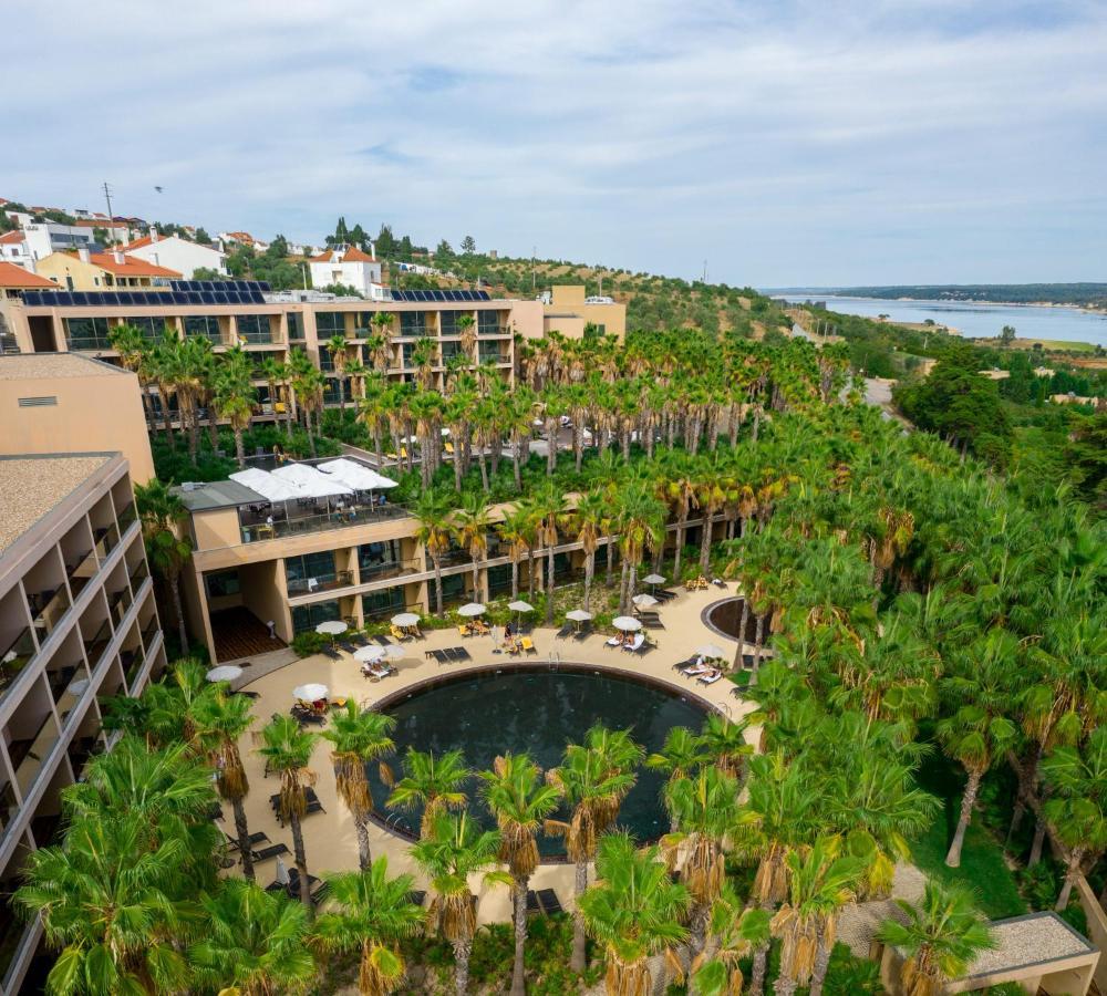 Ap Lago Montargil - Conference & Spa Εξωτερικό φωτογραφία The photo shows a modern hotel complex set amidst lush greenery, featuring numerous palm trees. In the foreground, there is a circular swimming pool surrounded by lounge chairs. The hotel itself has multiple levels with balconies overlooking the pool