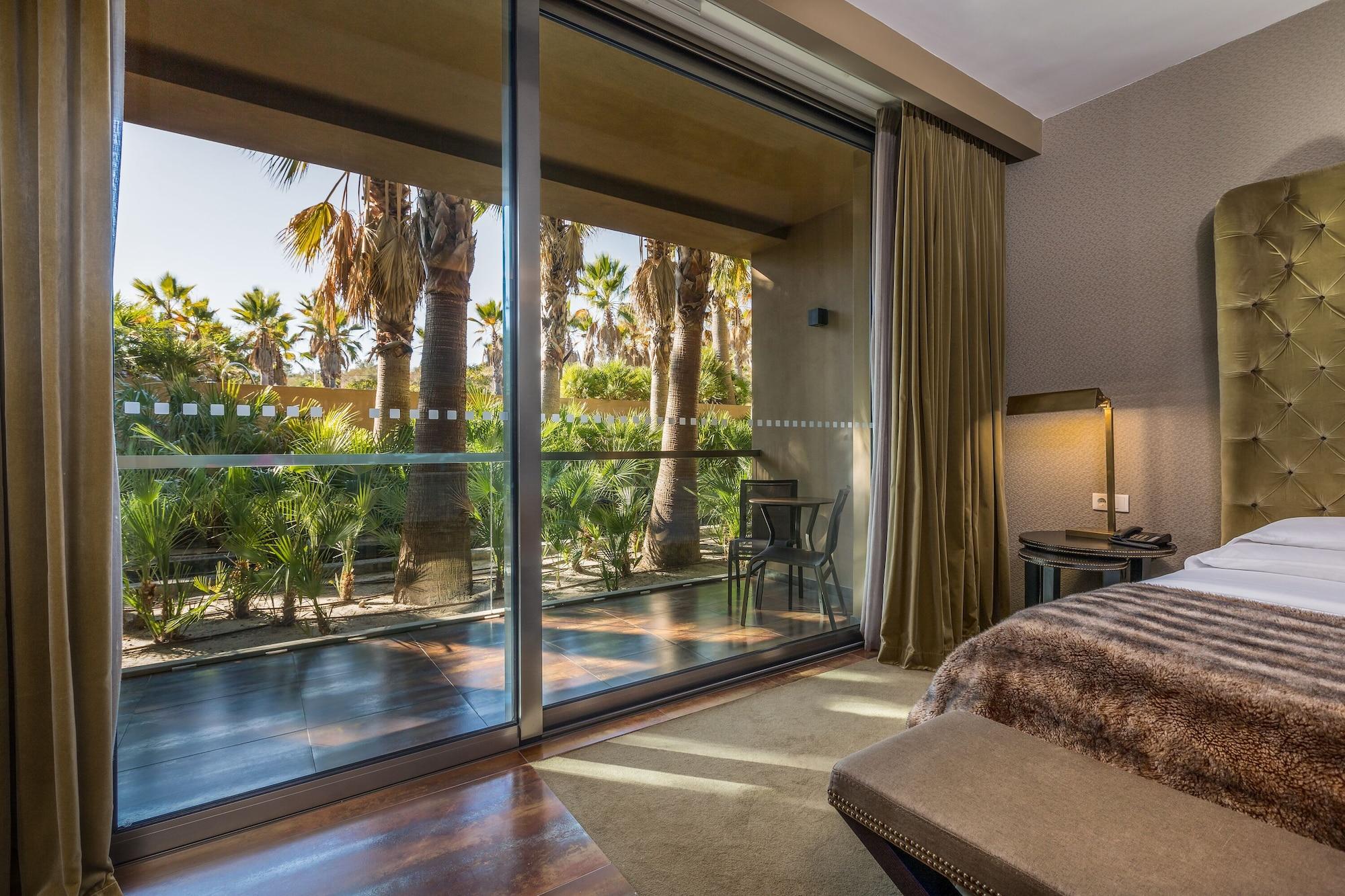Ap Lago Montargil - Conference & Spa Εξωτερικό φωτογραφία The photo shows a bedroom with large glass doors that lead out to a balcony or terrace. The view outside features palm trees and lush greenery, indicating a tropical or warm climate. Inside the room, there is a bed, a bedside table with a lamp, and a