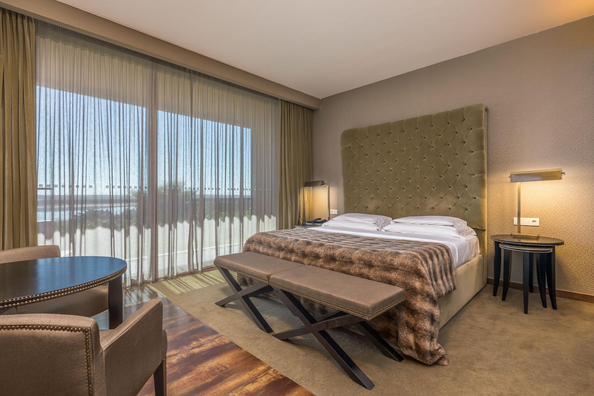 Ap Lago Montargil - Conference & Spa Εξωτερικό φωτογραφία The photo shows a modern hotel room featuring a large bed with a tufted headboard and a cozy blanket. There are curtains draped over large windows that allow natural light to flow in, providing a view of the outside. A small seating area includes two