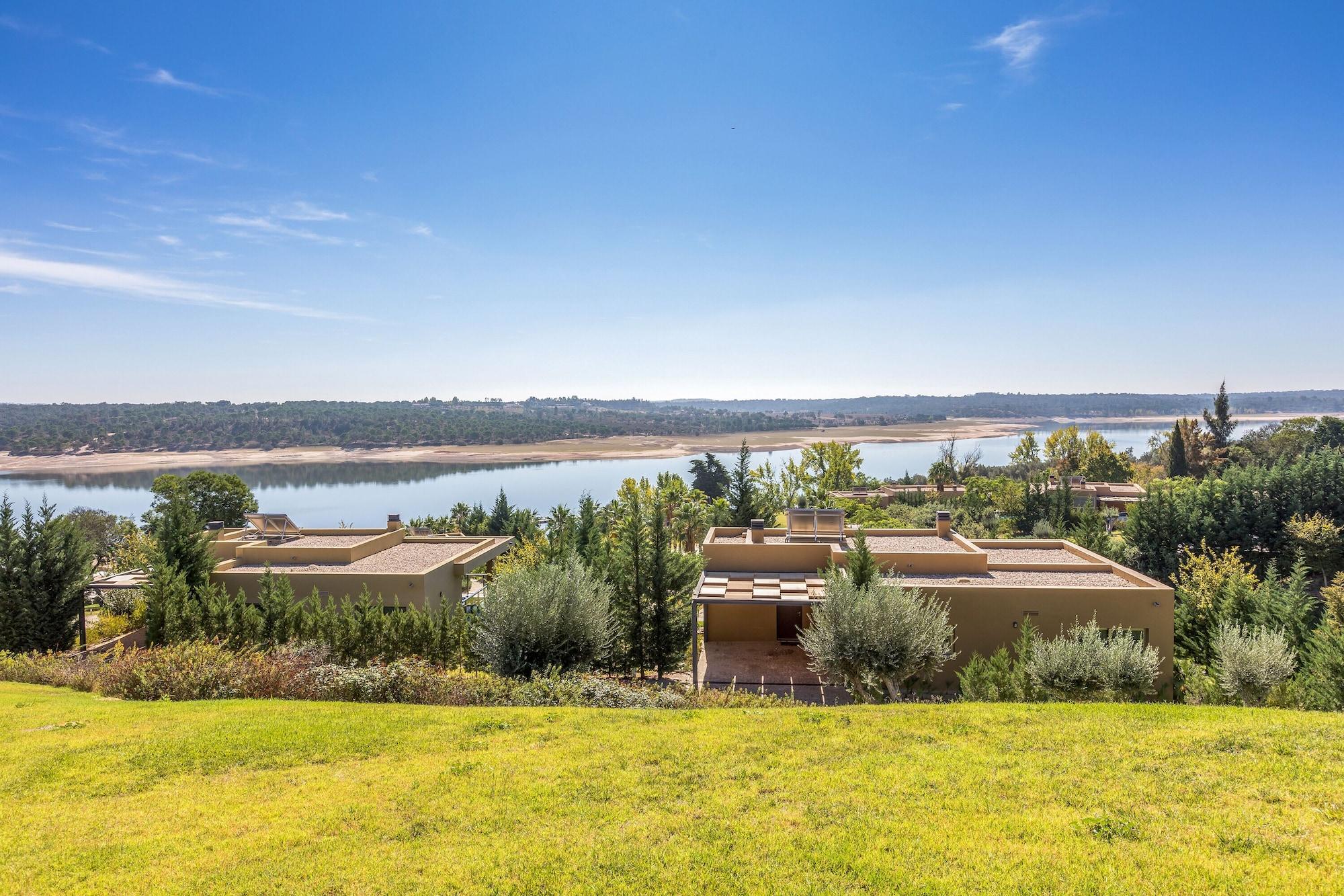 Ap Lago Montargil - Conference & Spa Εξωτερικό φωτογραφία The photo depicts a scenic landscape featuring a calm body of water, possibly a lake or river, surrounded by lush greenery and trees. In the foreground, there are several modern buildings with flat roofs, set against a vibrant blue sky. The overall s