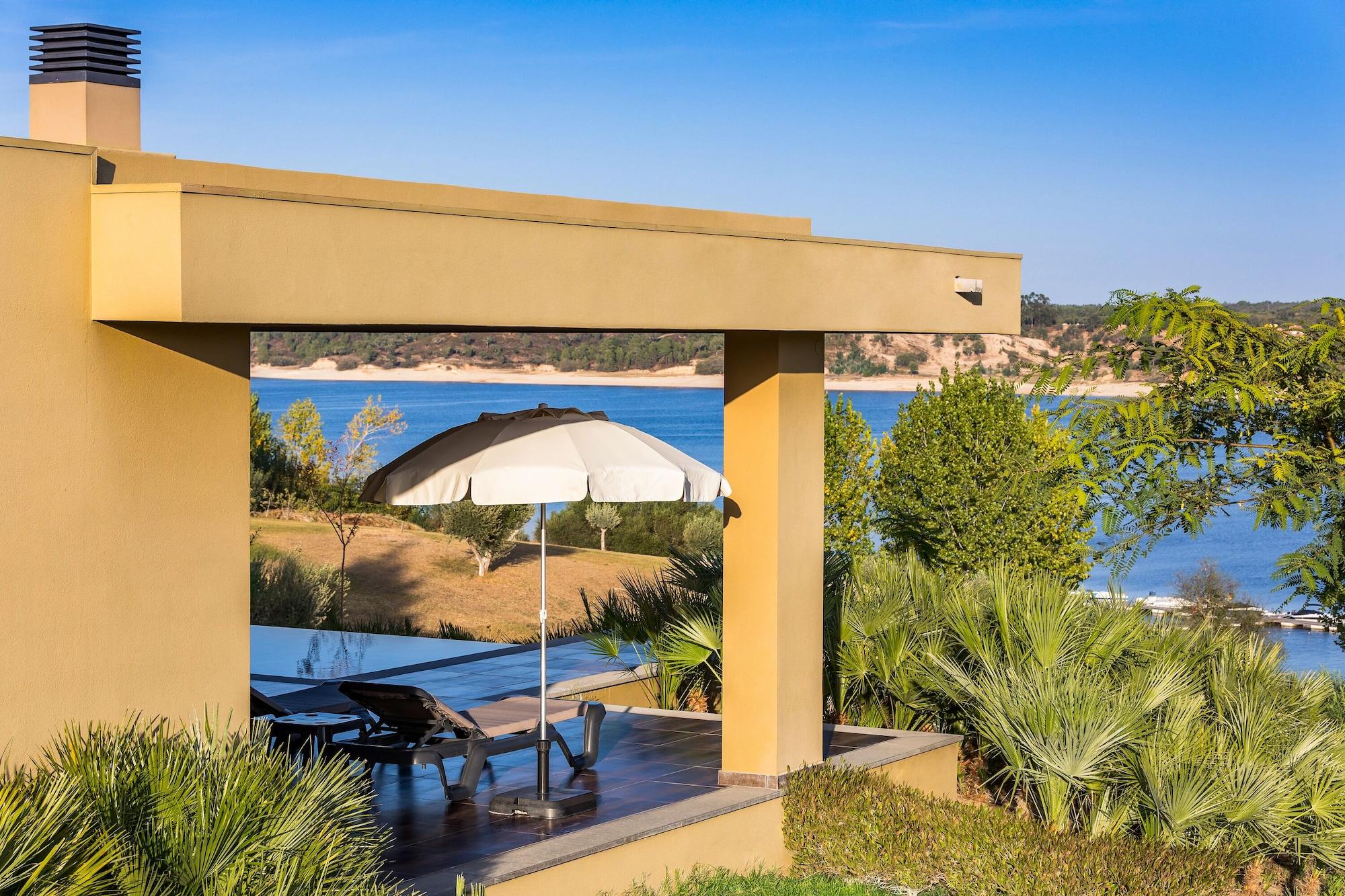 Ap Lago Montargil - Conference & Spa Εξωτερικό φωτογραφία The photo shows a peaceful outdoor area with a modern architectural structure featuring an overhang. There is a lounge chair positioned beneath a large umbrella, creating a shaded spot to relax. In the background, there is a picturesque view of a bod