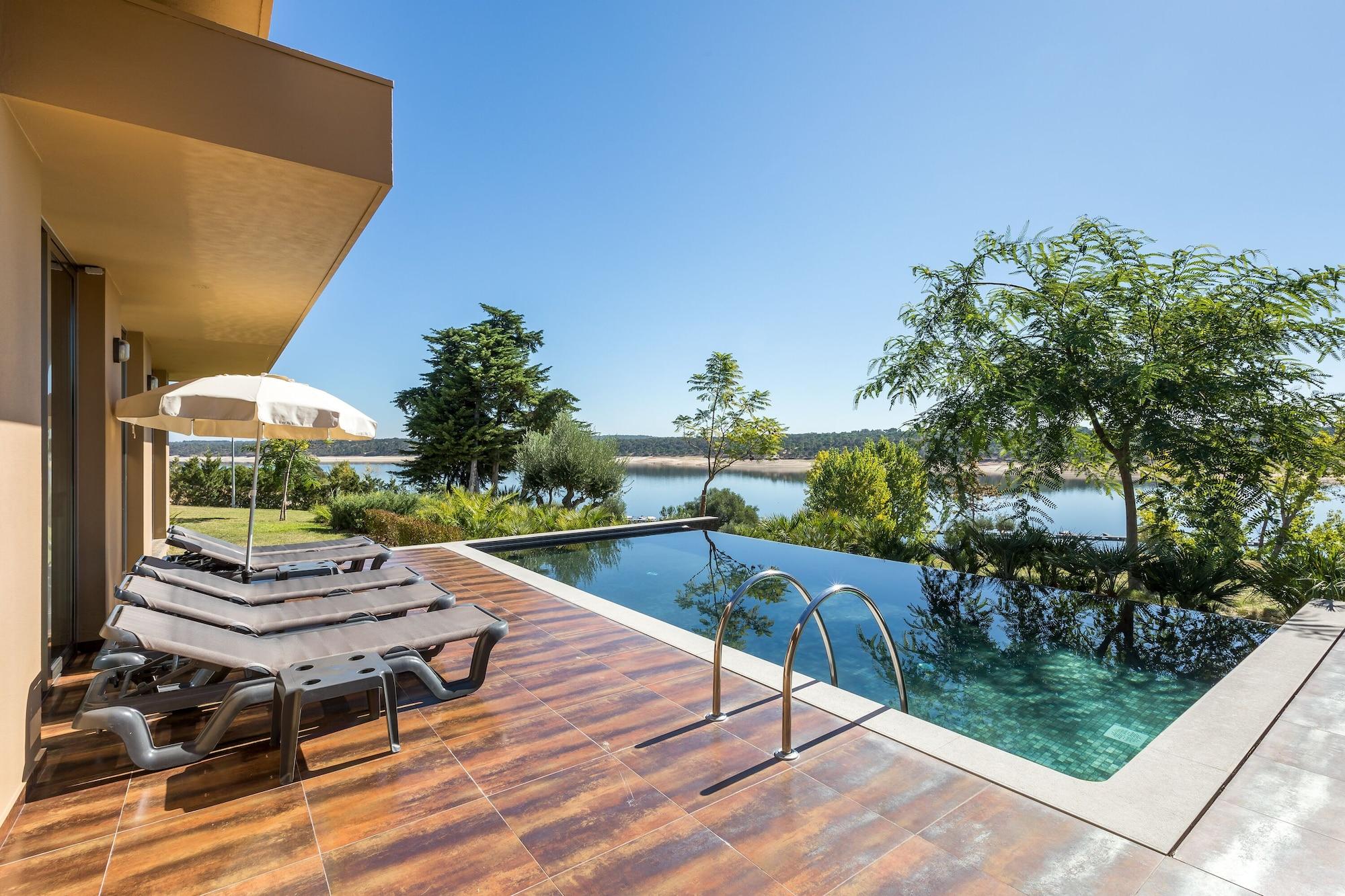 Ap Lago Montargil - Conference & Spa Εξωτερικό φωτογραφία The photo shows a luxurious outdoor setting by a pool. There are several lounge chairs arranged next to an infinity pool, which overlooks a scenic body of water and lush greenery. A parasol provides shade for relaxation. The sky is clear and blue, in