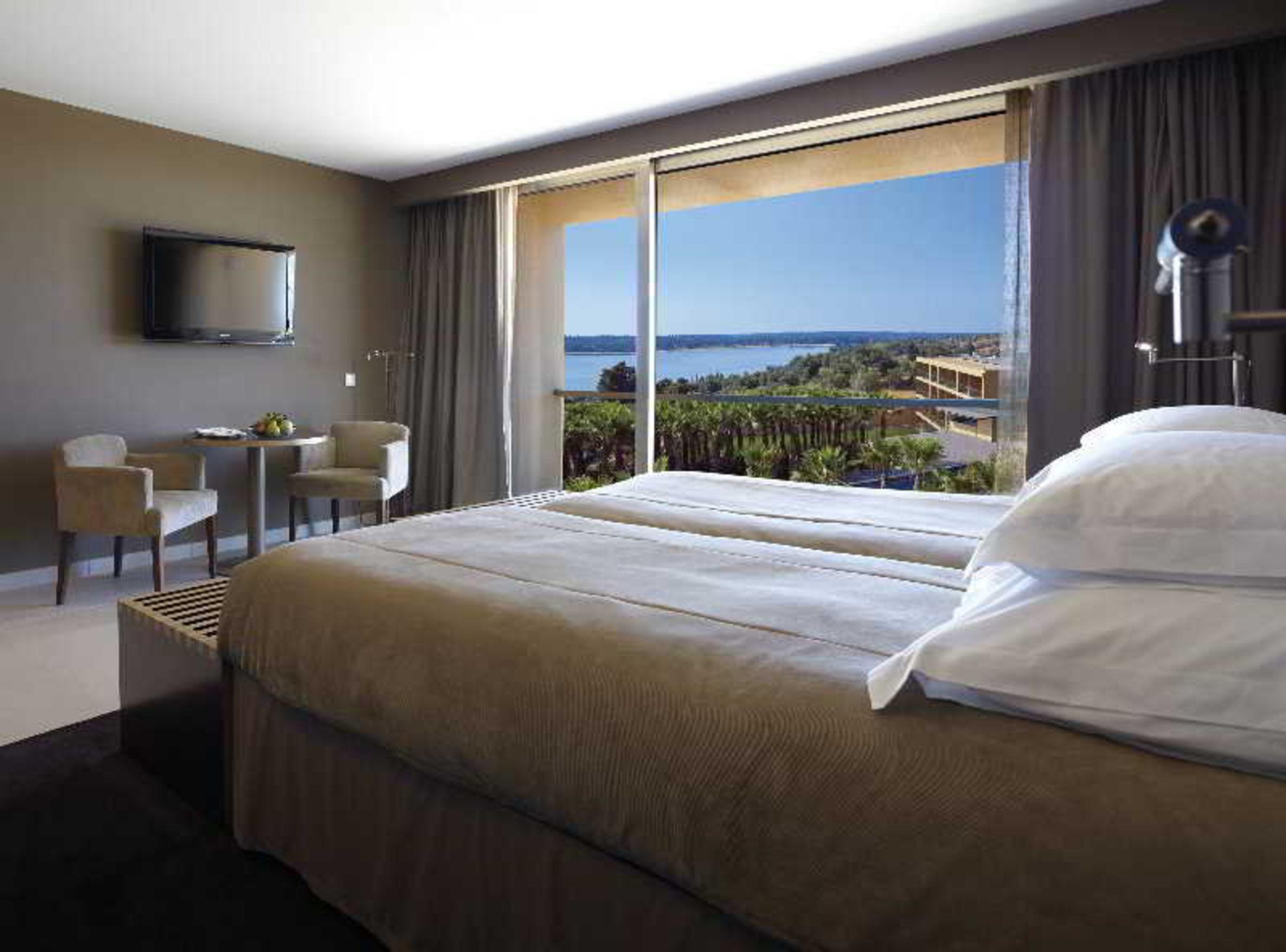 Ap Lago Montargil - Conference & Spa Εξωτερικό φωτογραφία The photo shows a modern hotel room with a large window offering a view of the sea and mountains. The room features a comfortable bed with white pillows and brown bedding. There is a small sitting area with two chairs and a table, as well as a wall-m