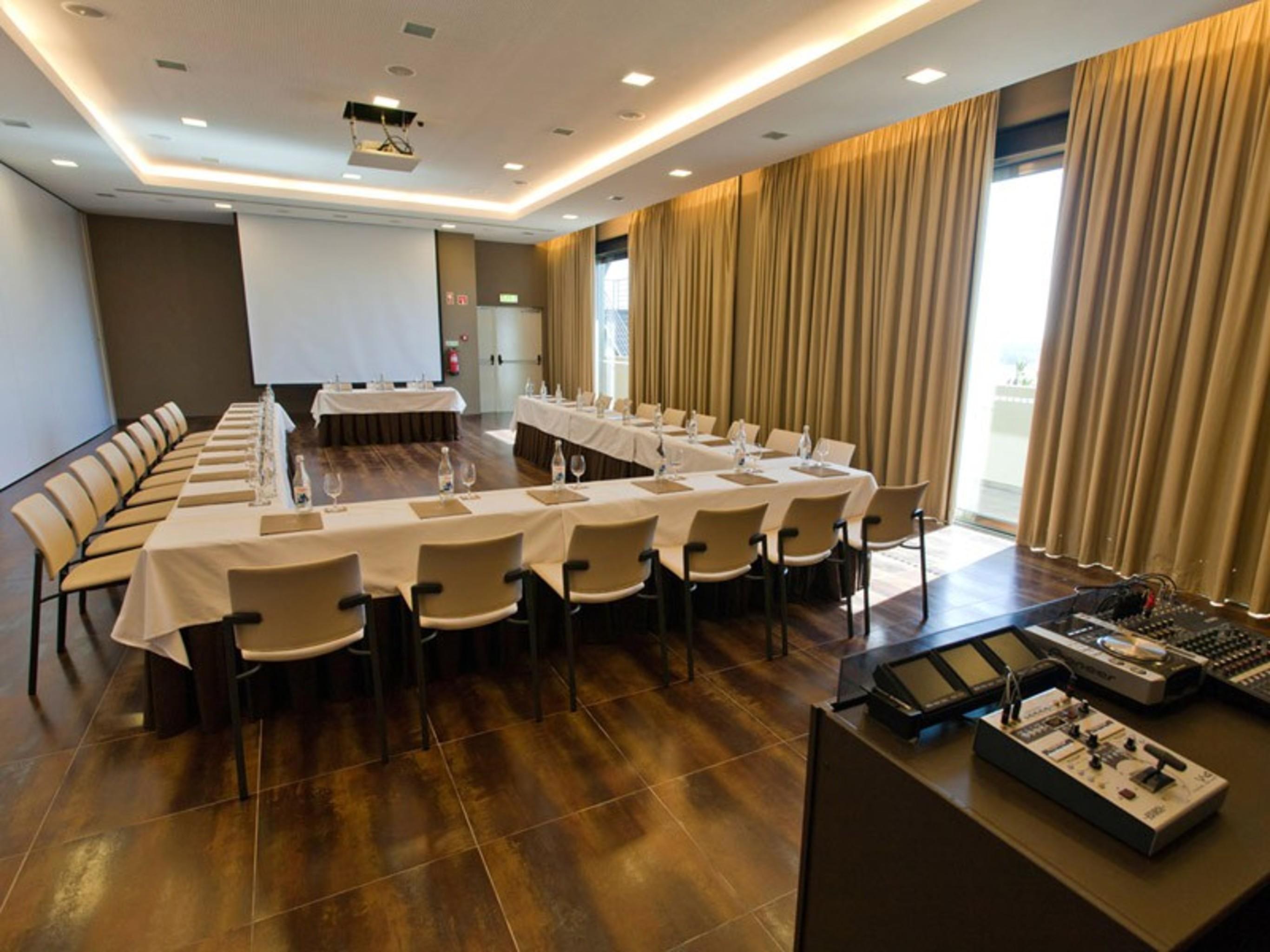 Ap Lago Montargil - Conference & Spa Εξωτερικό φωτογραφία The photo shows a modern conference room set up for a meeting. There are several tables arranged in a U-shape, surrounded by chairs. A large screen and projector are positioned at the front, with a table for speakers or presenters. The room has large