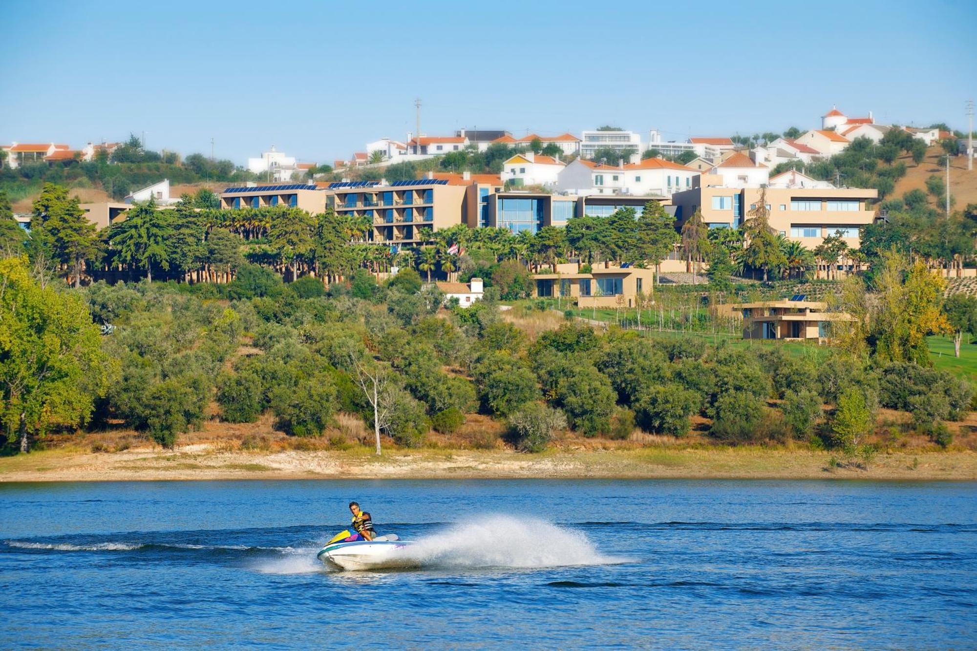 Ap Lago Montargil - Conference & Spa Εξωτερικό φωτογραφία The photo shows a person riding a jet ski on a body of water, creating a spray as they speed across the surface. In the background, there are modern buildings and a hillside with trees and greenery. The sky is clear and blue, suggesting a bright and 