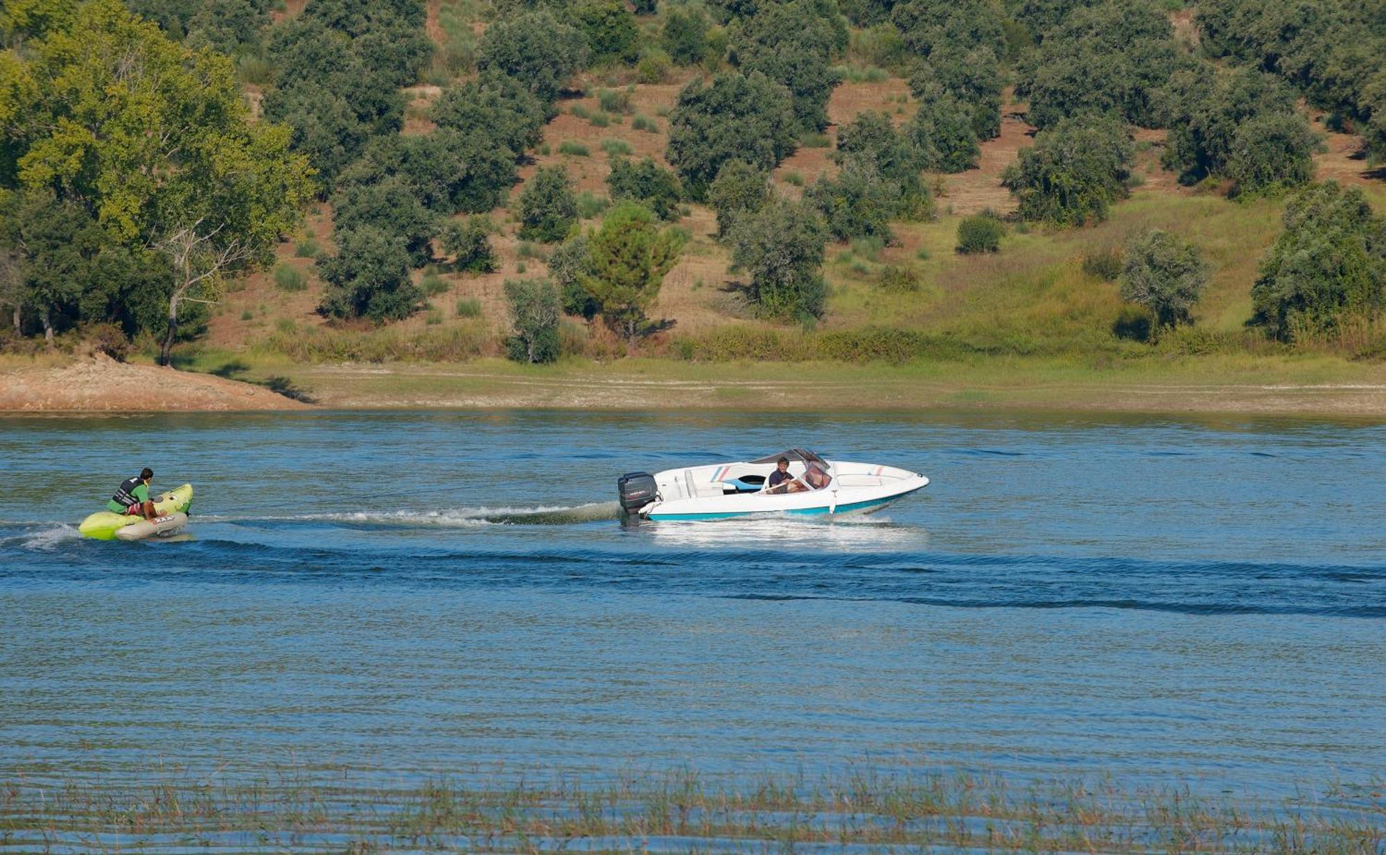 Ap Lago Montargil - Conference & Spa Εξωτερικό φωτογραφία The photo shows a scene on a water body, likely a river or lake. In the foreground, a speedboat is moving quickly through the water, leaving a wake behind it. There are two people visible in the boat. In the background, there is a hilly area covered 