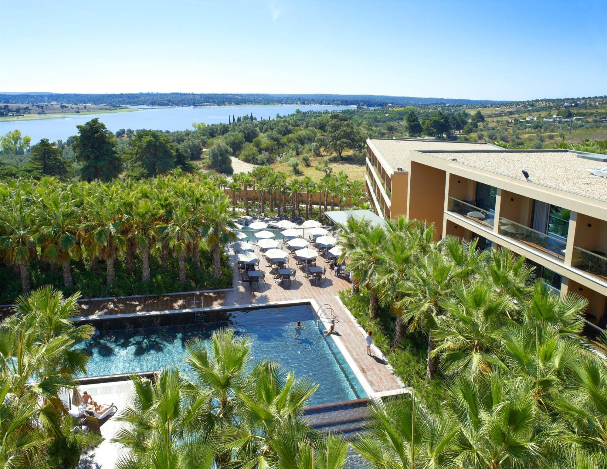 Ap Lago Montargil - Conference & Spa Εξωτερικό φωτογραφία The photo shows a scenic view of a modern hotel or resort. In the foreground, there is a swimming pool surrounded by palm trees, with lounge chairs positioned beside it. Sun umbrellas provide shade over the chairs. The background features a lush land
