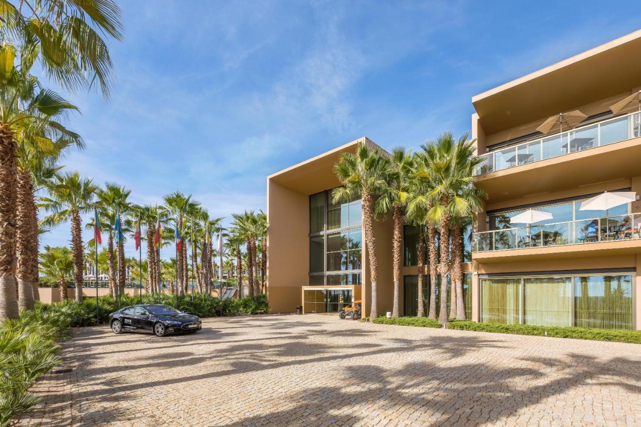 Ap Lago Montargil - Conference & Spa Εξωτερικό φωτογραφία The photo shows a modern building with a sleek design, featuring large glass windows and balconies. The structure is surrounded by a well-maintained outdoor area with palm trees, adding a tropical feel. In front of the building, there is a paved park